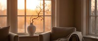 Cozy living room corner with armchair and decorative vase by large windows during golden hour sunset creating warm atmospheric lighting for home interior design.