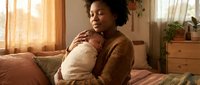 Young African American mother gently holding swaddled newborn baby in warm sunlit bedroom, eyes closed in peaceful moment of bonding.