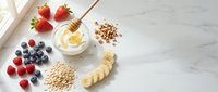 Healthy breakfast ingredients with fresh berries, banana slices, oats, nuts and honey on white marble surface for nutritious morning meal preparation.