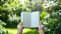 Hands holding open blank notebook against blurred green garden background with sunlight and bokeh effect, creating peaceful reading atmosphere outdoors.
