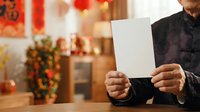 Man in black shirt holding blank white card with festive red and gold decorations blurred in background, suitable for holiday greeting cards, seasonal promotions, and celebration marketing materials.