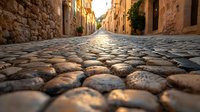 Cobblestone street perspective in Mediterranean town at sunset, with ancient stone walls and warm sunlight reflecting off rounded paving stones.