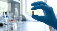 Scientist in blue gloves holding small glass vial with sample in modern laboratory setting with equipment and glassware for research and analysis.