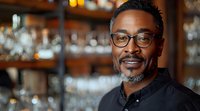 Confident African American male bartender with glasses and beard smiling in upscale bar with blurred bottles background. Professional portrait for hospitality industry.