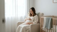 Pregnant woman in beige dress sitting in nursery chair touching belly, expecting mother in peaceful maternity moment preparing for baby arrival.