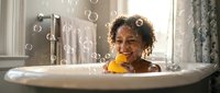 Young African American girl enjoying bubble bath with rubber duck toy in white bathtub. Child playing happily in soapy water with floating bubbles around bathroom.