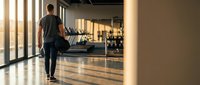 Young athletic man walking through modern fitness gym interior with large windows and exercise equipment in background during golden hour lighting.