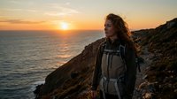 Woman hiker with backpack standing on rocky coastal cliff at golden hour sunset, gazing at ocean horizon during adventure travel and outdoor exploration.