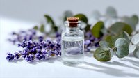 Small glass bottle with essential oil surrounded by fresh lavender flowers and eucalyptus leaves on white fabric, ideal for aromatherapy and wellness.