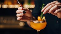 Professional bartender garnishing orange cocktail with fresh rosemary sprig in elegant coupe glass at upscale bar with warm ambient lighting.