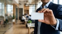Mature male professional with dark skin tone wearing navy business suit holding blank white card in modern office environment with colleagues working in background.