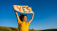 Young boy in yellow shirt holding yes flag outdoors on sunny day with clear blue sky and green field, expressing positivity and affirmation.