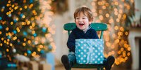 Excited young boy laughing with Christmas gift box, sitting on turquoise chair against blurred holiday lights and decorated tree background.