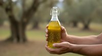 Hand holding glass bottle of golden olive oil with orchard trees blurred in background, suitable for food, cooking, agriculture, and culinary marketing materials.