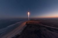 Rocket launch at dusk over coastal landscape with sandy beach and ocean, creating dramatic light trail against darkening sky.