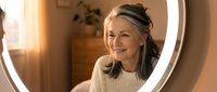 Mature woman with gray hair smiling at reflection in round mirror during morning skincare routine in cozy bedroom with warm natural lighting.