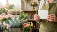 Florist in olive apron holding blank white card in flower shop with pink blooms and greenery, suitable for business cards, floral services, and small business marketing.