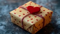 Gift box wrapped in kraft paper with red heart pattern, tied with red and white twine string, decorated with felt heart on top, against dark background.