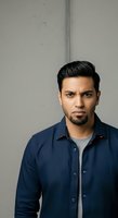 Young Middle Eastern man with dark hair and beard wearing navy button-up shirt over gray undershirt, posed against neutral gray wall background for portrait and professional use.