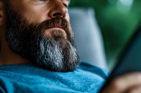 Close up of mature Caucasian man with full dark beard wearing blue shirt relaxing at home, partial face view showing contemplative expression against blurred background.