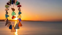 Colorful sea glass wind chime hanging against golden sunset over calm ocean water creating peaceful meditation and relaxation atmosphere for home decor.
