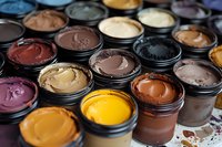 Colorful collection of professional makeup foundation creams in open containers showing various skin tone shades from light to dark, arranged in rows for cosmetic artistry.