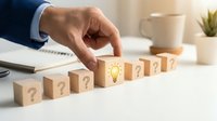Businessman hand selecting wooden cube with light bulb icon among question mark blocks on office desk representing creative problem solving and innovation.