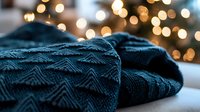 Cozy knitted dark teal blanket with chevron pattern texture against blurred warm Christmas lights bokeh background creating festive winter atmosphere.