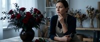 Thoughtful woman sitting at kitchen table with red roses bouquet, contemplating by window in cozy home interior during quiet morning moment.