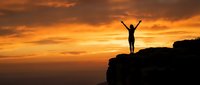 Silhouette of person with raised arms celebrating success on mountain cliff edge against dramatic orange sunset sky for motivational content.
