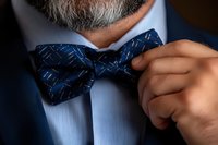 Mature businessman adjusting elegant navy blue bow tie with geometric pattern on formal white dress shirt for special occasion or event.