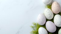 Speckled pastel Easter eggs in soft pink, mint and white shades arranged with delicate green foliage on white marble background with copy space.