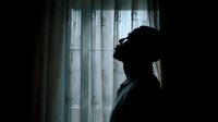Silhouette of person leaning against wall near window with sheer curtains, conveying solitude and contemplation in dramatic lighting.