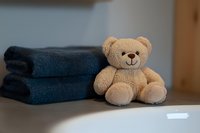 Soft plush teddy bear sitting next to folded navy blue towels on gray surface, creating cozy bathroom or spa atmosphere with warm lighting and shallow depth of field.