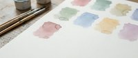 Watercolor paint palette with soft pastel color swatches on textured paper, art supplies and brushes visible, creative workspace for artistic projects.