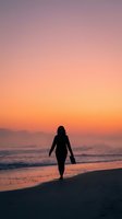Silhouetted female surfer in wetsuit walking toward ocean waves at golden hour sunset on sandy beach with dramatic sky.