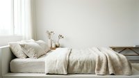 Cozy bedroom interior with unmade bed, cream linen bedding, decorative pillows, dried flowers in vase on nightstand, natural window light, minimalist Scandinavian style home decor.