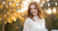 Young Caucasian woman with long wavy hair in white lace dress smiling outdoors with warm golden bokeh lights background during sunset hour for lifestyle photography.