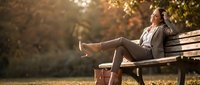 Young mixed-race businesswoman relaxing on park bench in autumn, listening to music with headphones, legs up, golden fall foliage background.