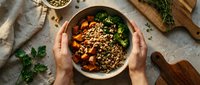 Hands holding bowl of roasted vegetables with quinoa and fresh herbs on kitchen counter, overhead view for food styling and culinary content.