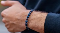 Male wrist wearing dark blue lapis lazuli natural stone beaded bracelet with silver accent beads against black sweater sleeve background.