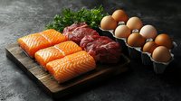 Fresh raw salmon fillets, beef steaks, and brown eggs arranged on rustic wooden board against dark background for protein-rich meal preparation.