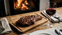 Grilled steak on wooden cutting board with red wine glass, fireplace background, fine dining table setting for restaurant or food photography.