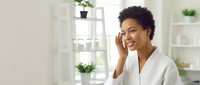 Young African American woman with curly dark hair wearing white blouse, smiling while touching face in bright modern office with shelving and plants behind her.