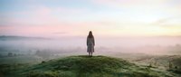 Woman standing alone on misty hillside at dawn, wearing long coat, gazing at foggy valley landscape below during peaceful morning sunrise.
