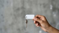 Person of medium skin tone holding a white keychain with a blank tag and silver key against a textured gray concrete wall background for real estate, property management, or moving concepts.