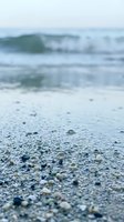 Close-up of sandy beach with shells and pebbles in foreground, blurred ocean waves and coastline in background. Shallow depth of field creates focus on sand texture for travel and nature designs.