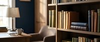 Wooden bookshelf filled with vintage books in a cozy home library, warm sunlight streaming through window, desk and lamp in background.