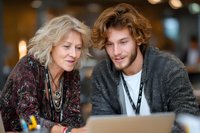 Senior woman and young man with curly hair looking at laptop together in cafe, sharing knowledge across generations.