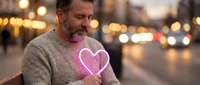 Middle-aged Caucasian man holding glowing pink neon heart shape outdoors at night with bokeh city lights background for romantic Valentine's Day concept.